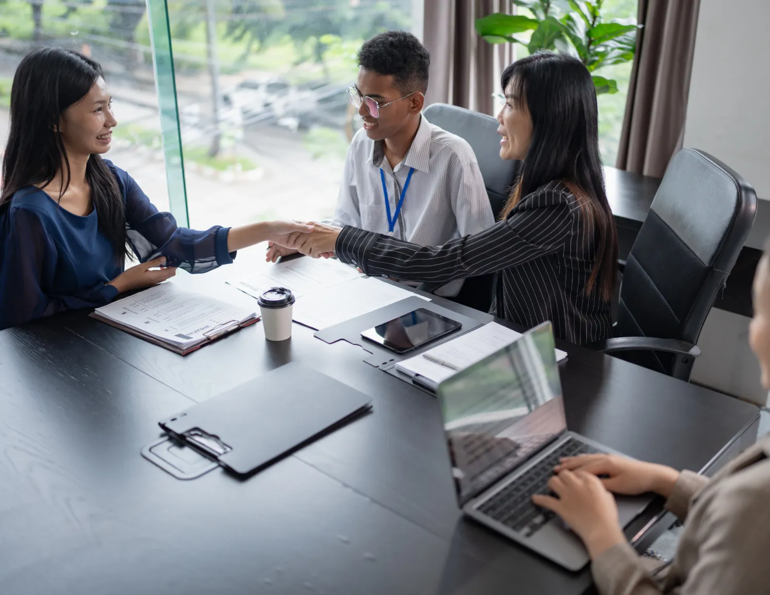 Sollicitatiegesprek van 4 mensen aan een tafel, schudden elkaar de hand, laptop en papier